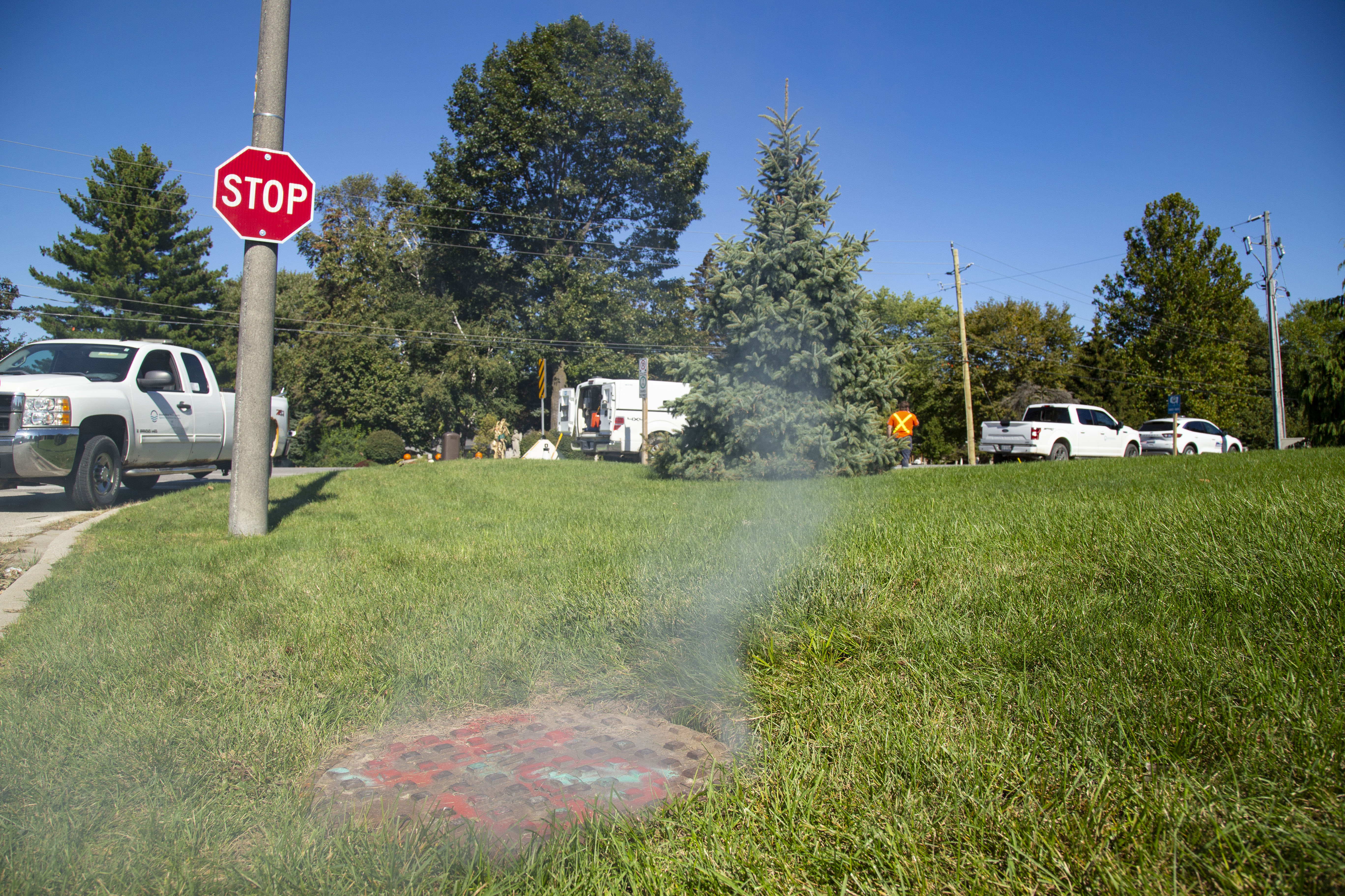 Sewer manhole with white vapour coming out of the holes. The manhole is on a grass lawn of a corner lot. Stop sign at the top left of image and work trucks in the background.