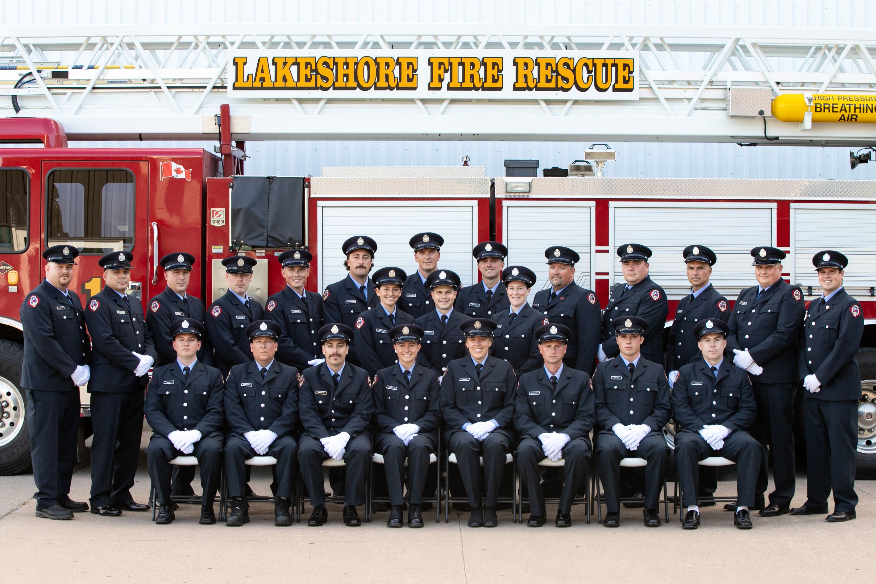 Group photo of the 2019 and 2022 fire recruit graduates. Firefighters in dress uniforms standing in front of a firetruck.