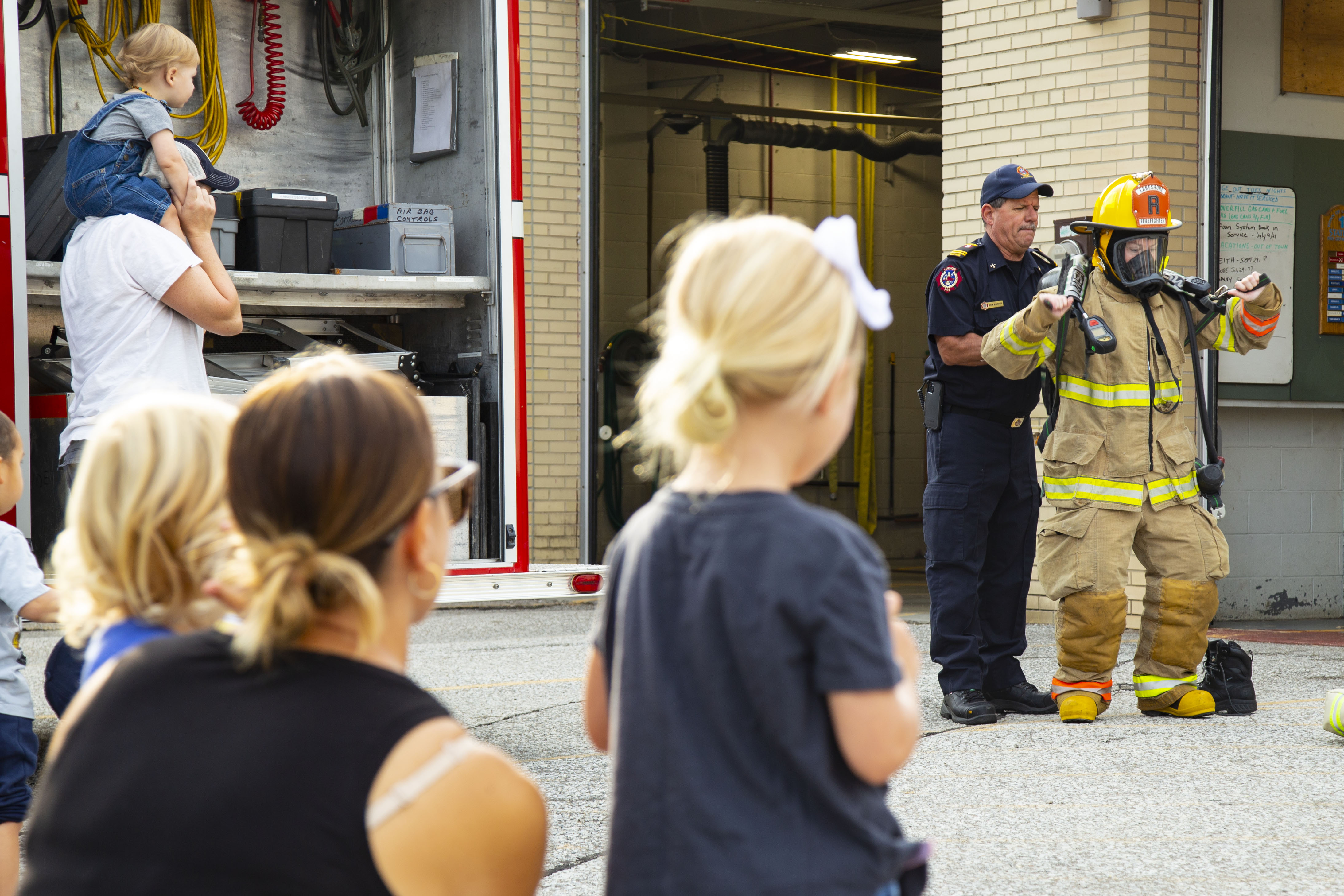 Crowd of people watching a firefighter don their protective bunker gear while another firefighter explains the process.