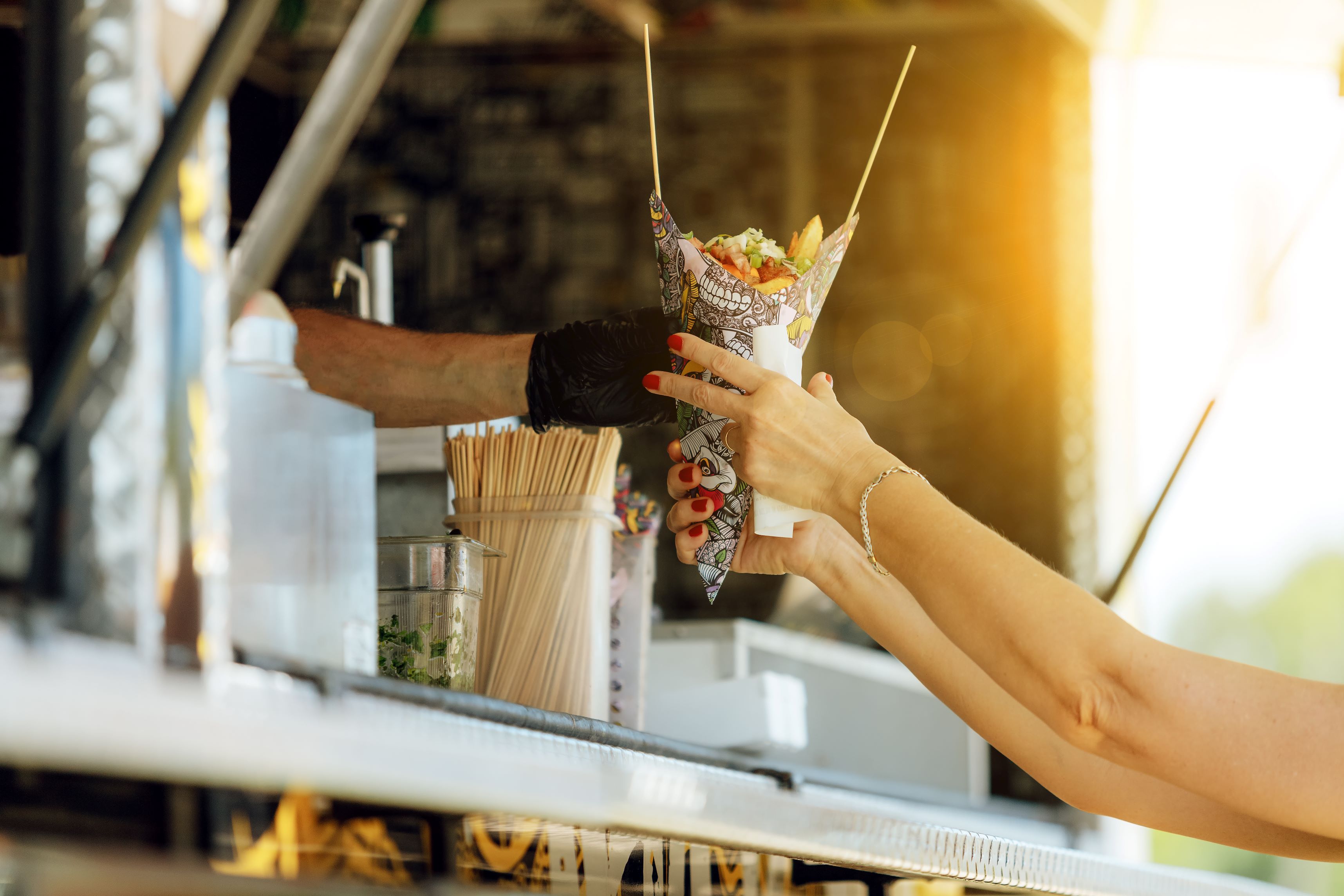 Photo of a gloved hand giving food to another person at a food truck with a sun flare in the background.