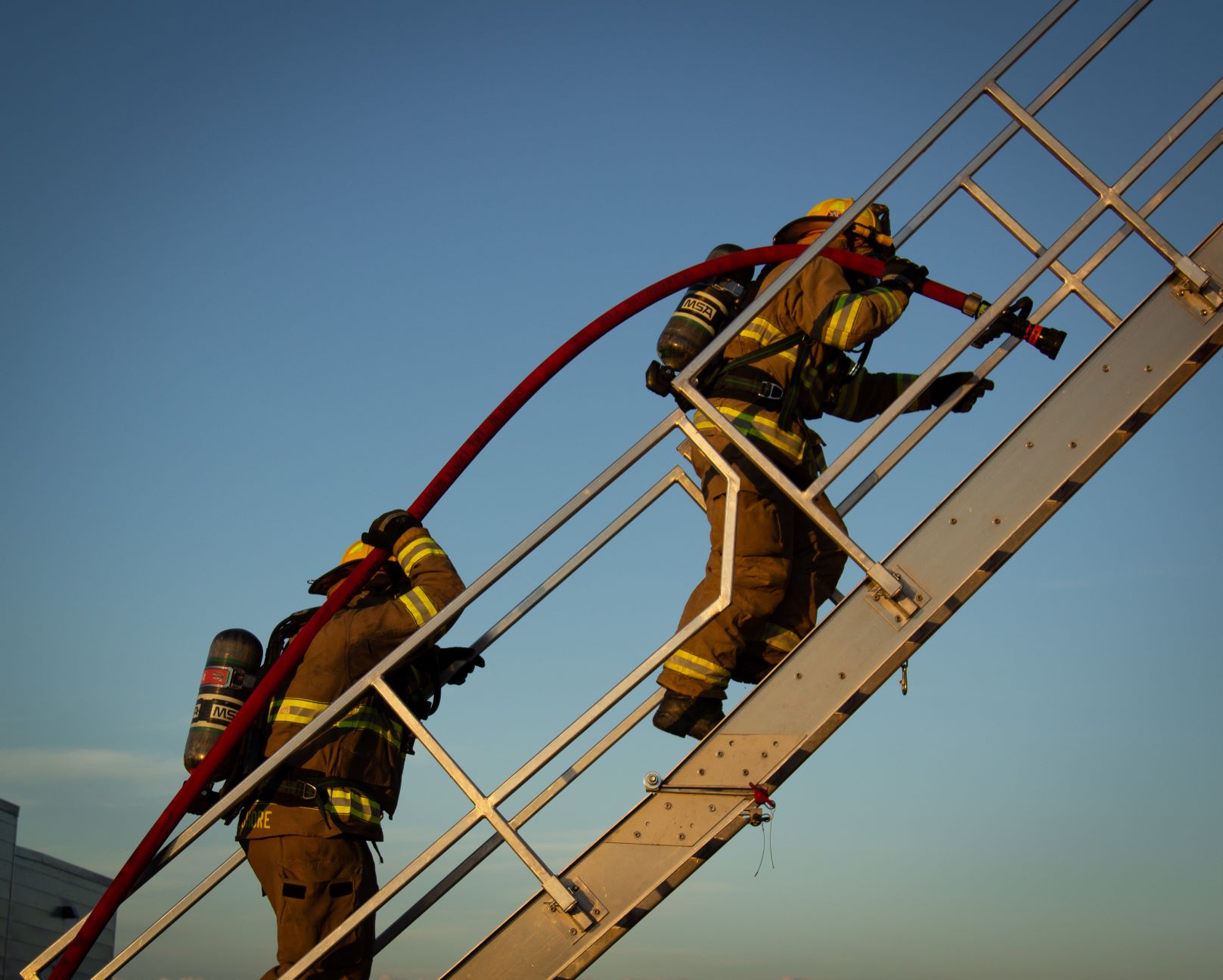 Photo of two firefighters climbing a ladder truck carrying a hose.