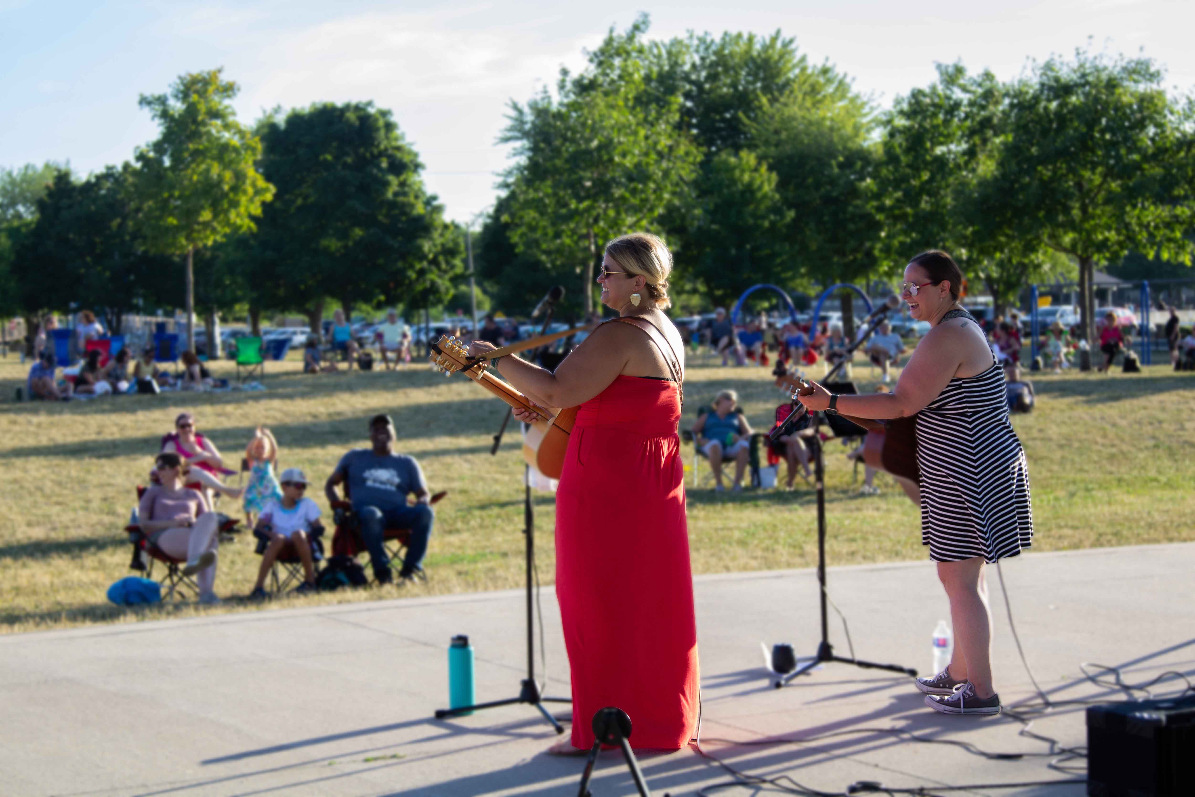 Photo of two people playing guitar and singing in front of a crowd of people sitting on the grass in a park.