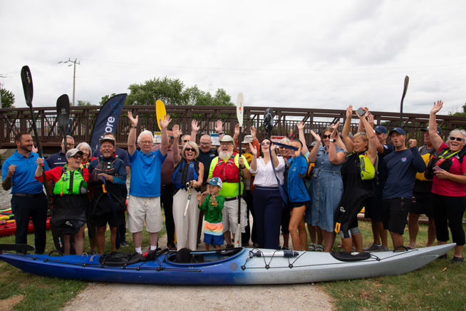 Group photo of council and community members with Kayak in front. Everyone is celebrating the opening of the new accessible kayak and canoe launch.